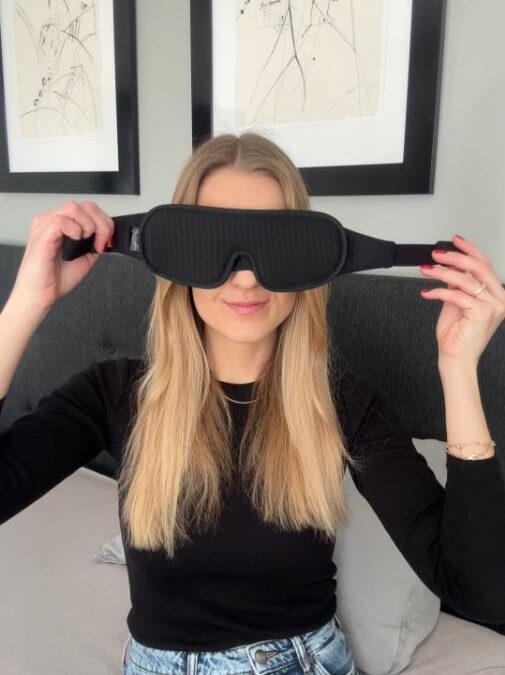 Kvinde med langt blondt hår holder sort sovemaske foran ansigtet i soveværelse