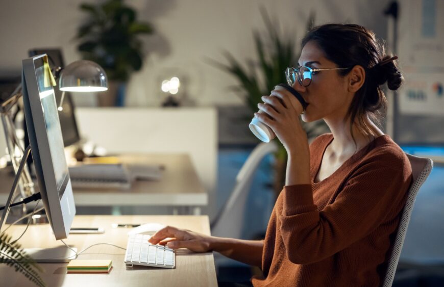 Ung kvinde med briller drikker takeaway-kaffe og arbejder ved computer på kontor om aftenen