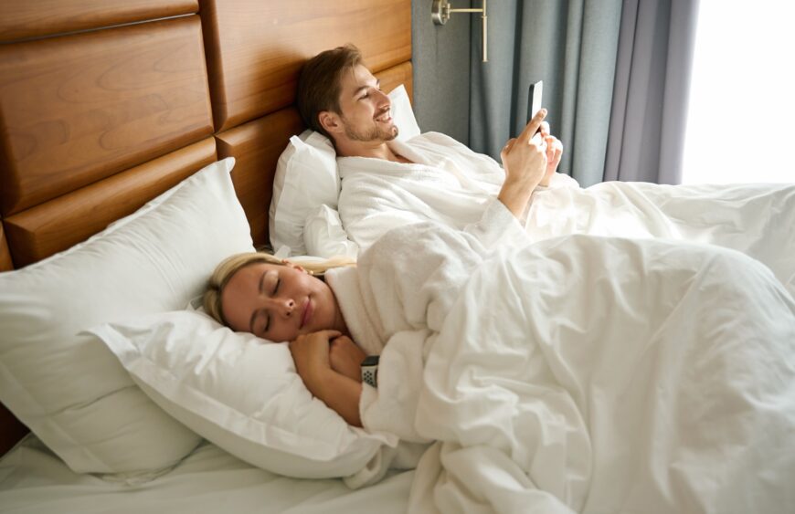 Mand i badekåbe ligger i stor seng og tjekker mobil, kvinde sover ved siden af under hvidt dyne