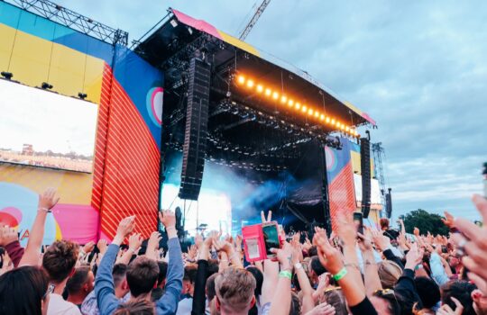 Stor udendørs musikfestivalscene under aftenhimmel, publikum med hænder i vejret