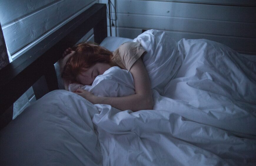 Person med rødt hår sover indsvøbt i hvidt dyne på mørk trærammeseng i dæmpet blåt lys