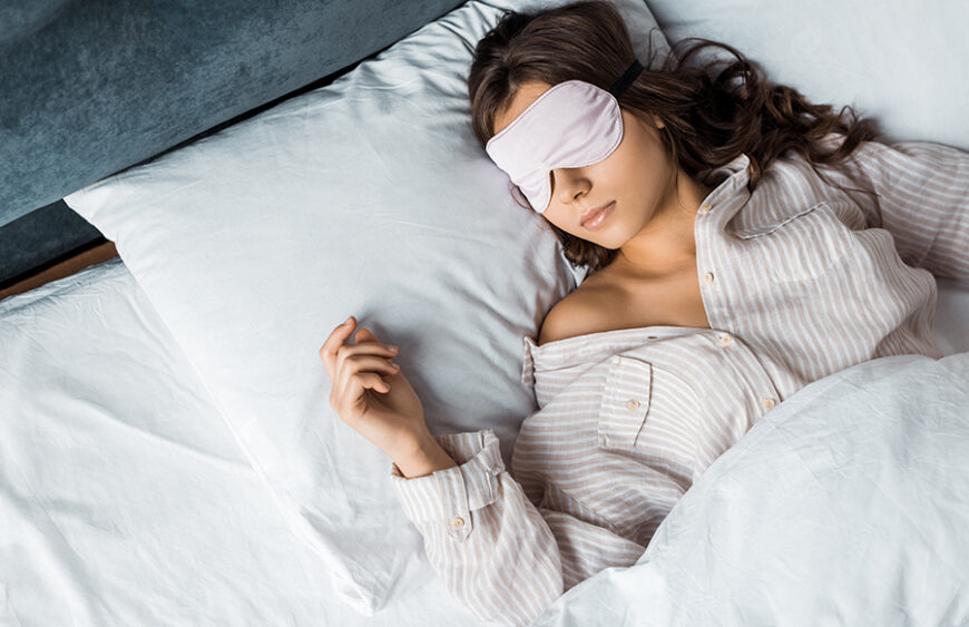 Ung kvinde med sovemaske og pyjamas sover roligt i hvidt sengetøj