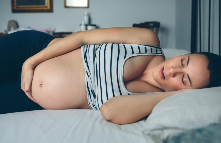 Gravid kvinde sover på siden i seng med hånd på maven, iført stribet top