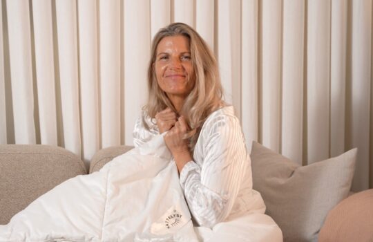 Smilende kvinde i hvid pyjamas holder blød dyne på sofa foran beige gardiner