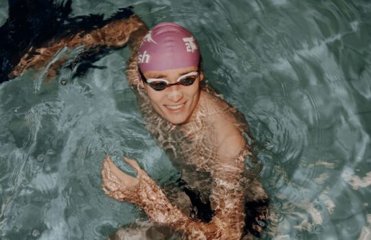Smilende svømmer med lilla badehætte og svømmebriller i pool, vandreflekser over arm og skuldre